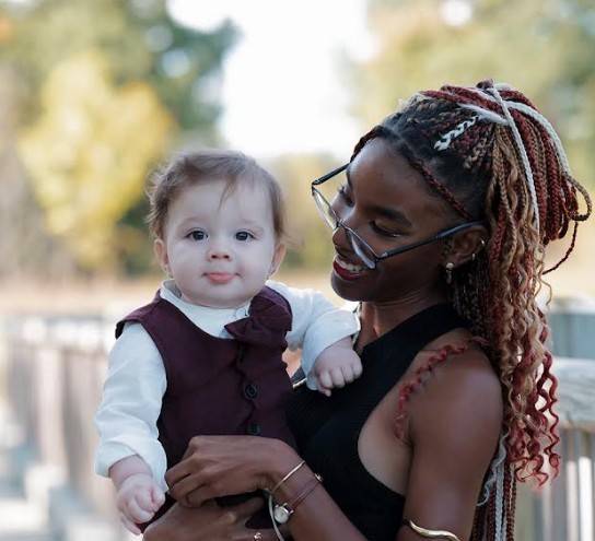 A woman with braided hair and glasses holds a baby wearing a burgundy outfit, both standing outdoors with blurred trees in the background.
