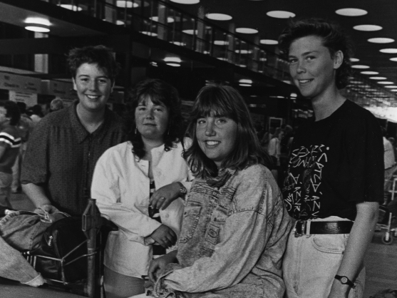 Four people pose together in an indoor setting with suitcases, dressed casually and smiling.