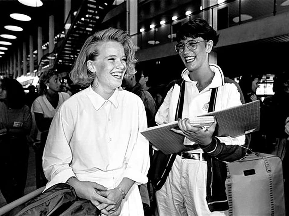 Two young adults laughing in an indoor setting, holding binders. The scene is lively, with many people in the background.
