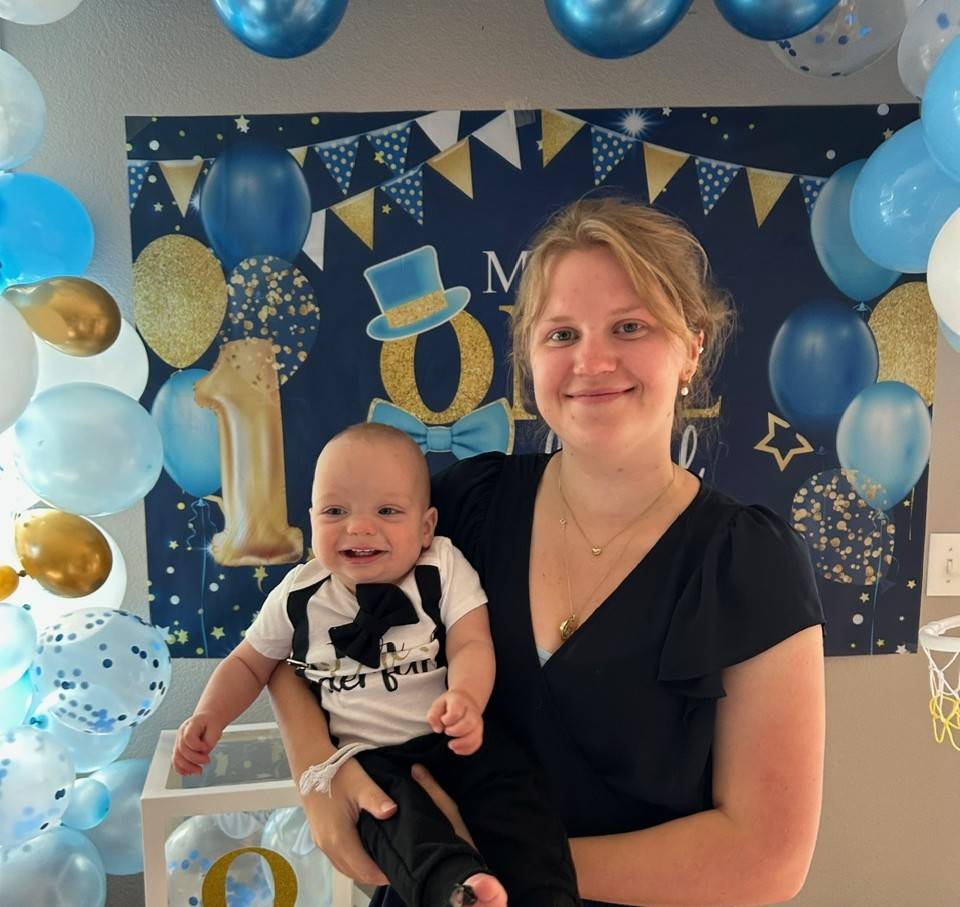 Woman holding a smiling baby at a first birthday party, surrounded by blue and gold balloons and festive decorations.