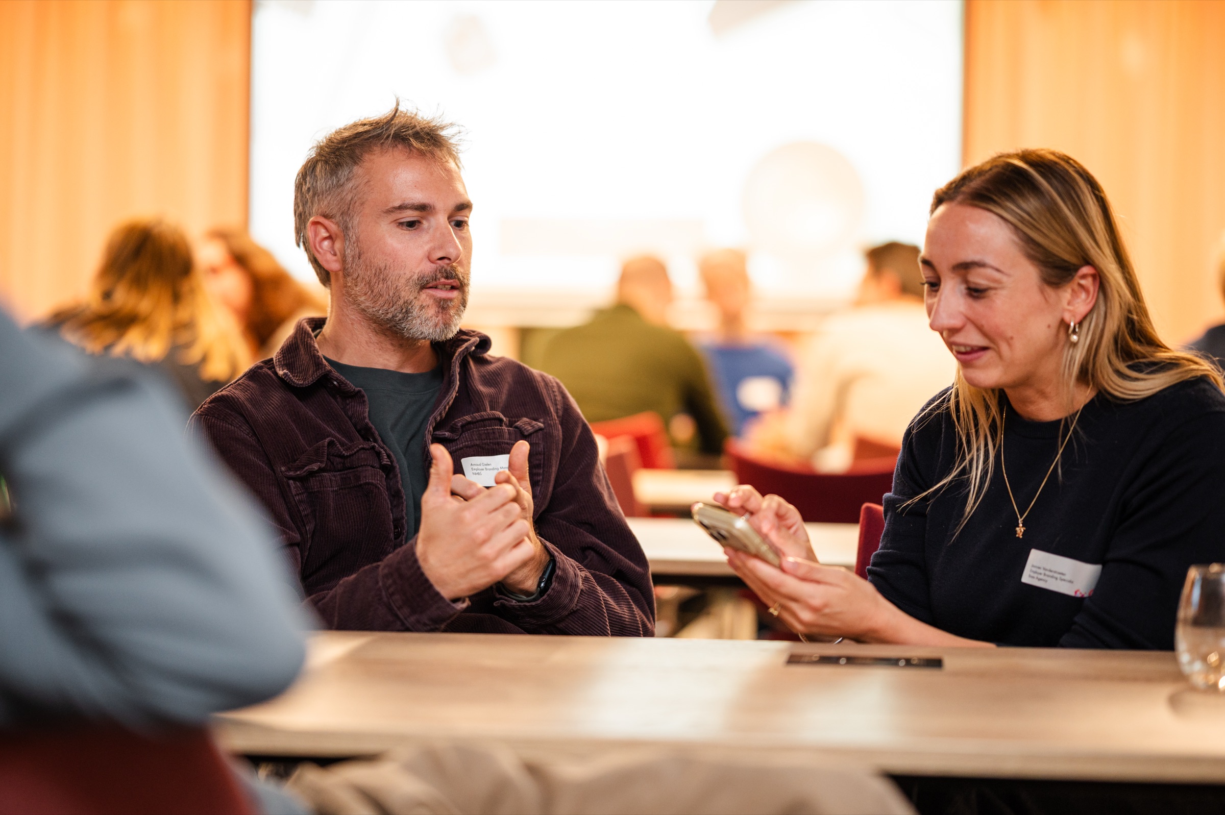 Two attendees in conversation at the round table, one of them showing something on a phone.