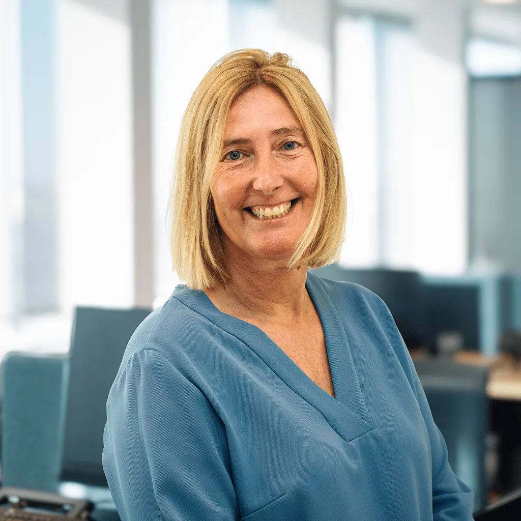 A smiling woman in an office environment, sitting at her desk with a computer and various office items.