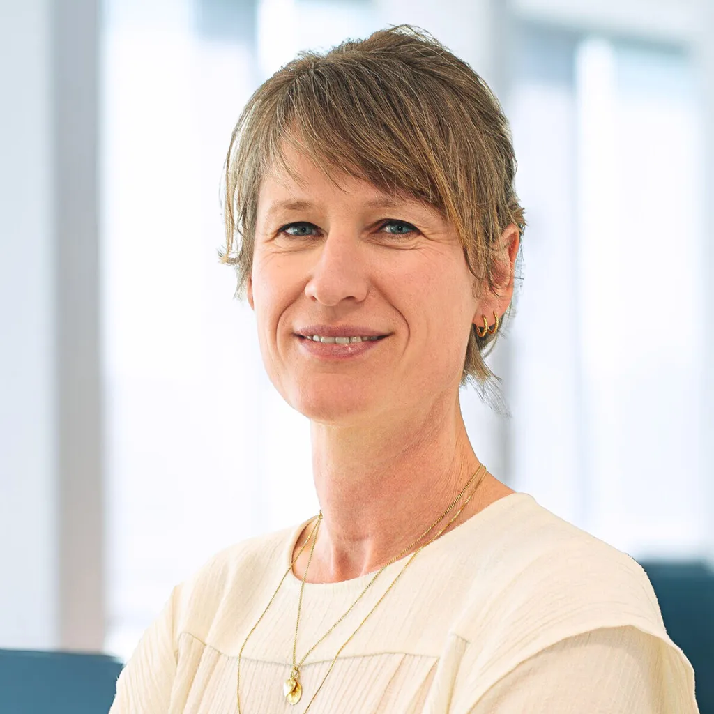 Smiling middle‑aged woman with short light brown hair in a cream top, wearing gold jewelry, posed in a bright office setting.