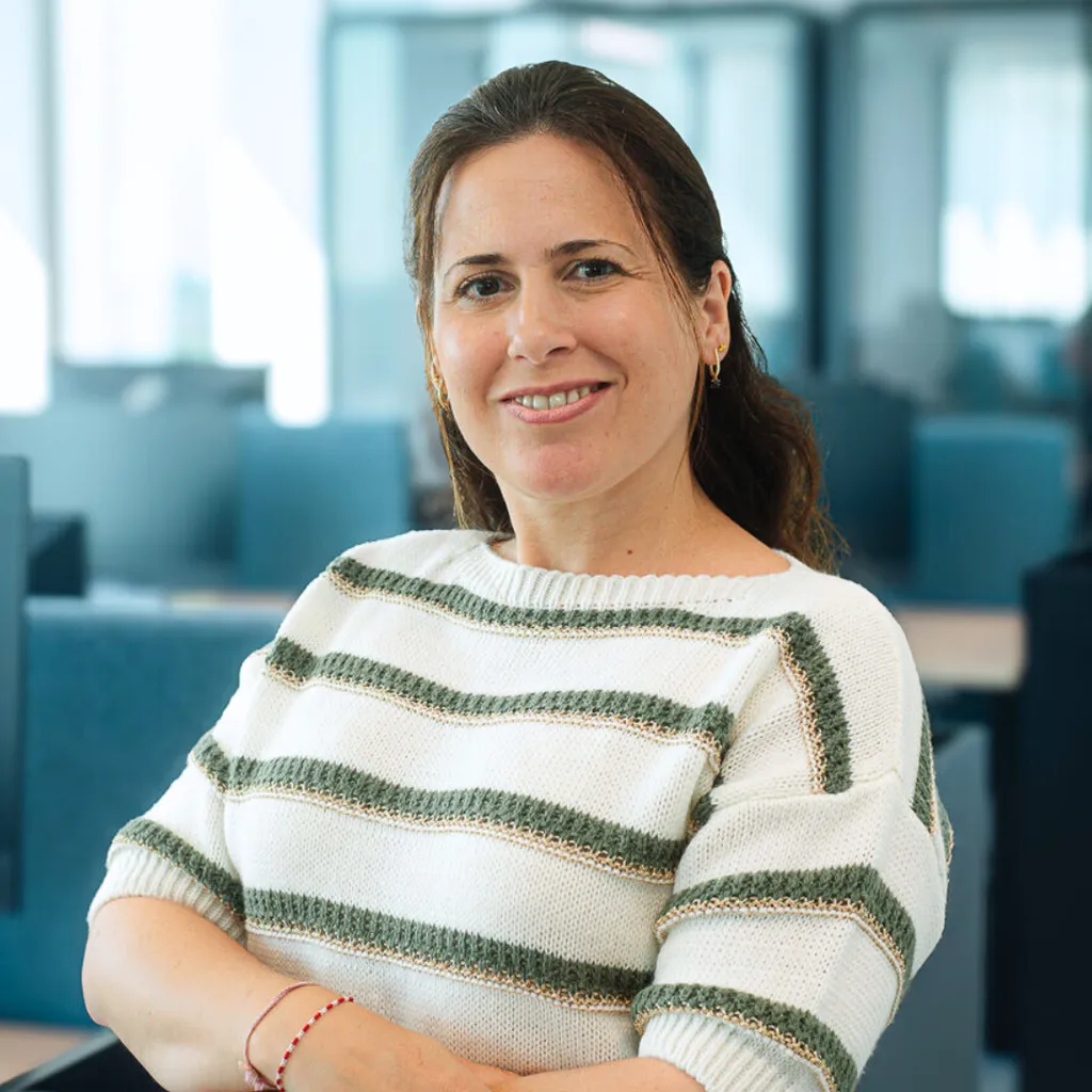 Smiling woman in a white sweater with green stripes, arms crossed, seated in a modern open-plan office.