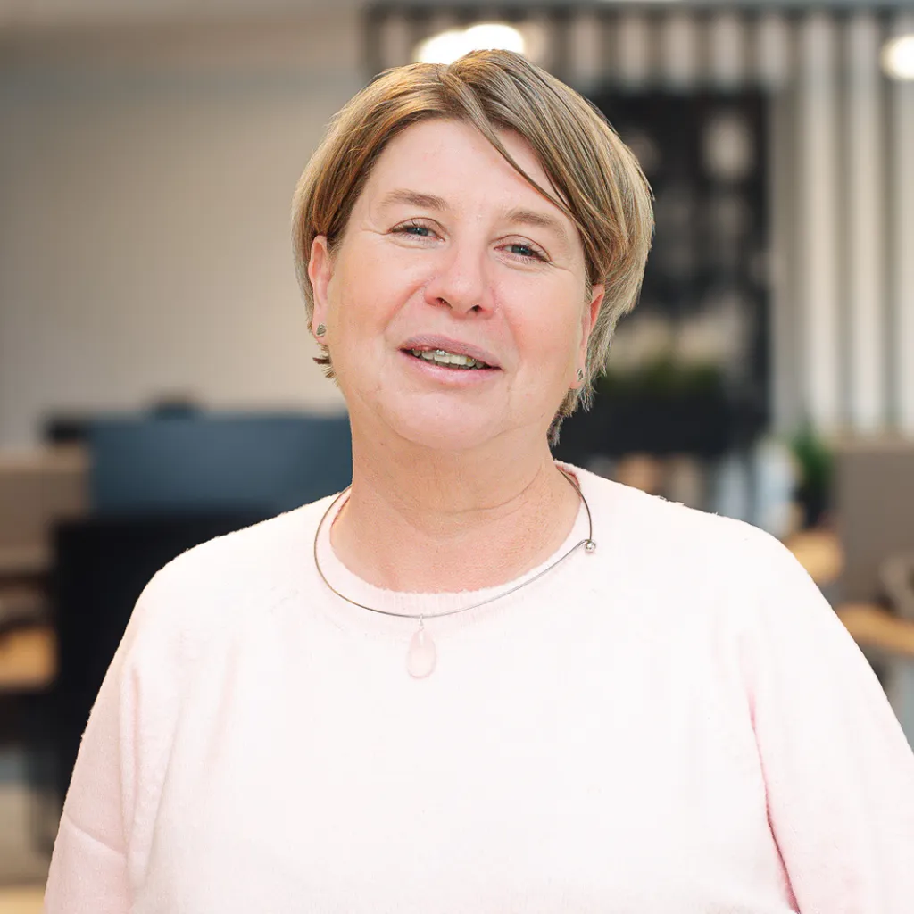 A person with short hair, wearing a light pink sweater, smiles in a blurred office setting.