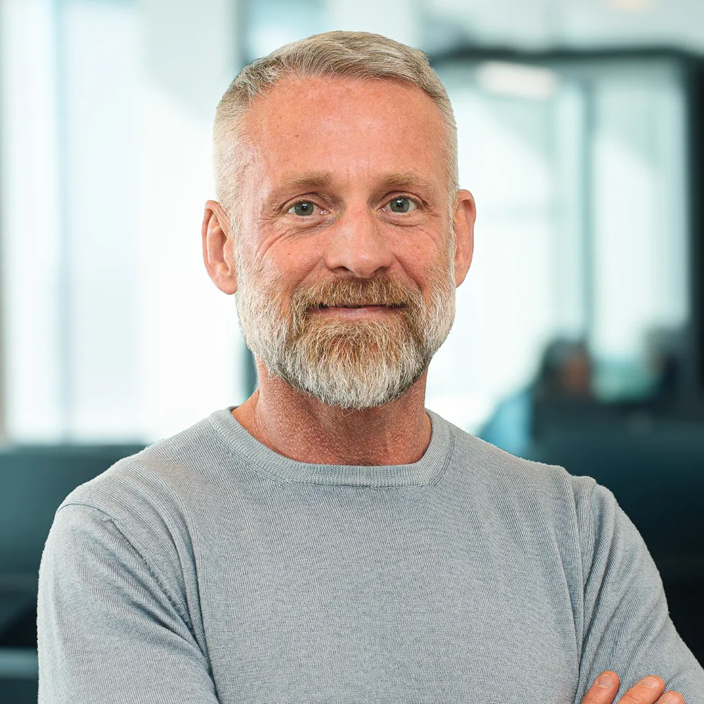 Middle‑aged man with gray hair and beard smiling with arms crossed in a bright modern office.