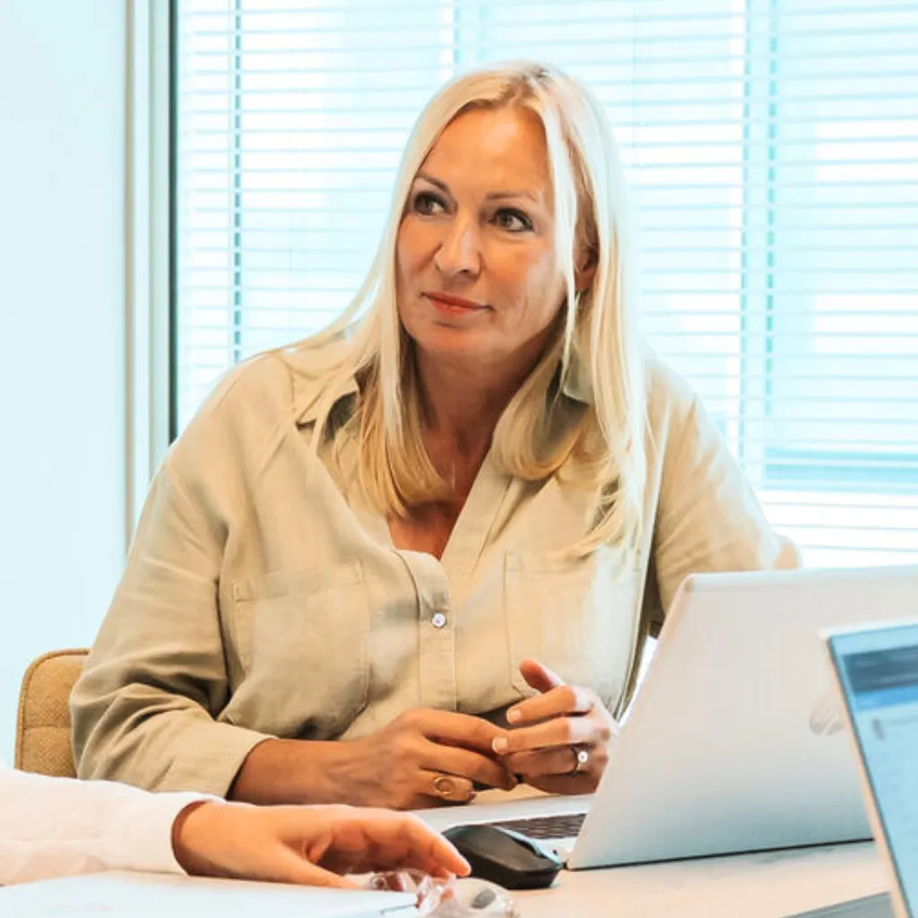 Blonde woman in beige shirt sits at a desk with a laptop, listening during a meeting in a bright office.