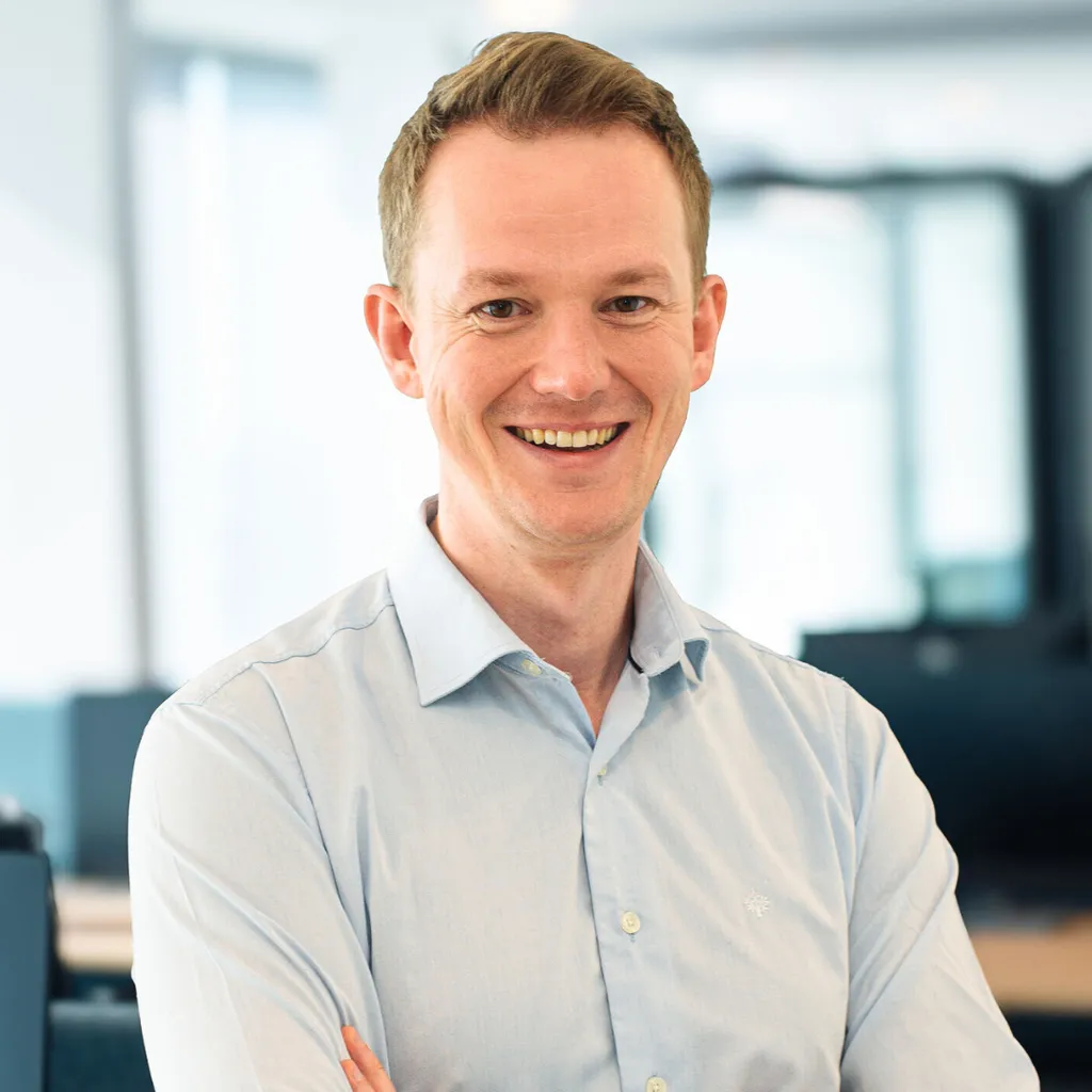 Smiling man in a light blue shirt stands with arms crossed in a bright office setting.