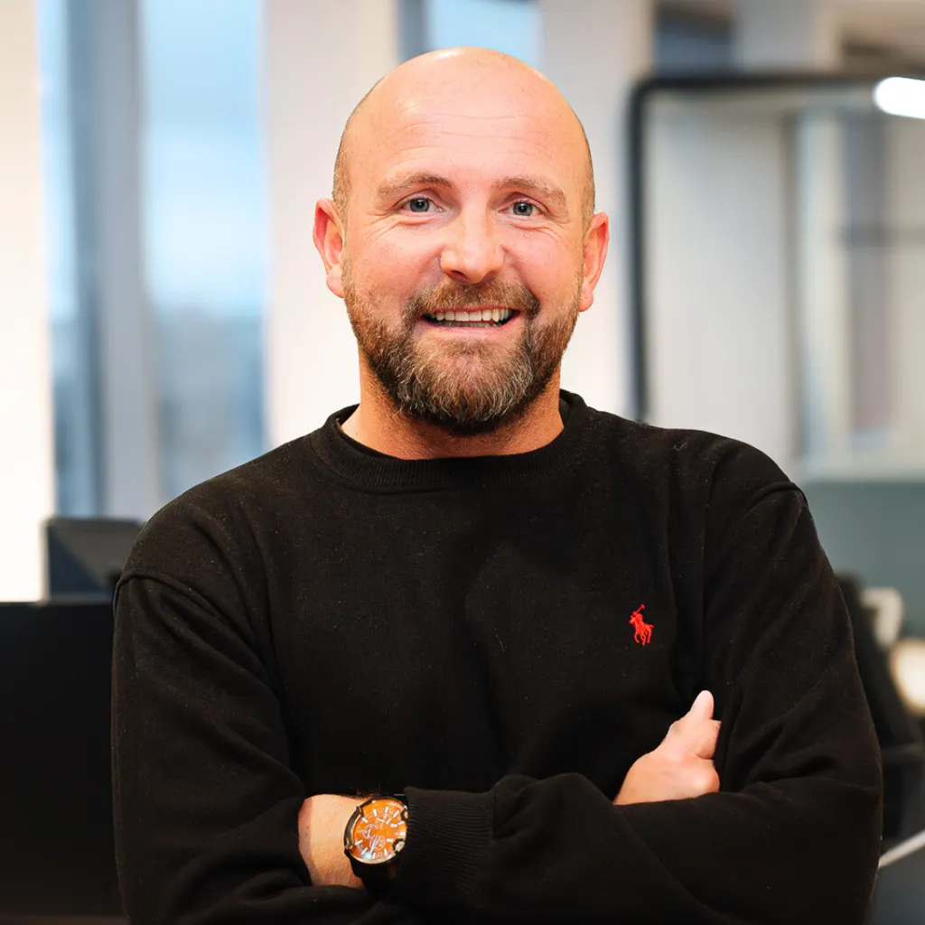 A smiling man with a beard, wearing a black sweater with a red logo, stands with arms crossed in a modern office setting.