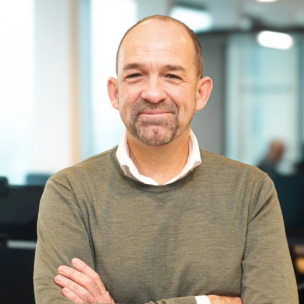 A man with a beard and mustache, wearing a green sweater and white shirt, stands with arms crossed in a bright office setting.