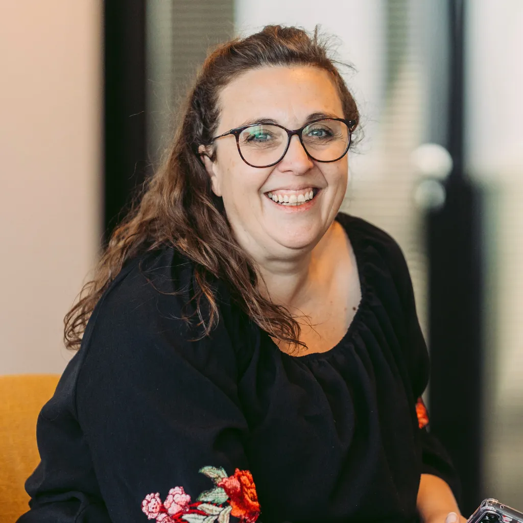 Smiling woman with glasses in a black top with floral sleeves, sitting indoors on a yellow chair, looking at the camera.
