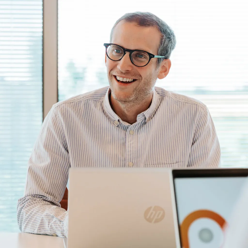 Smiling man with glasses and striped shirt sitting at a table with an HP laptop, in a bright room with blinds in the background.
