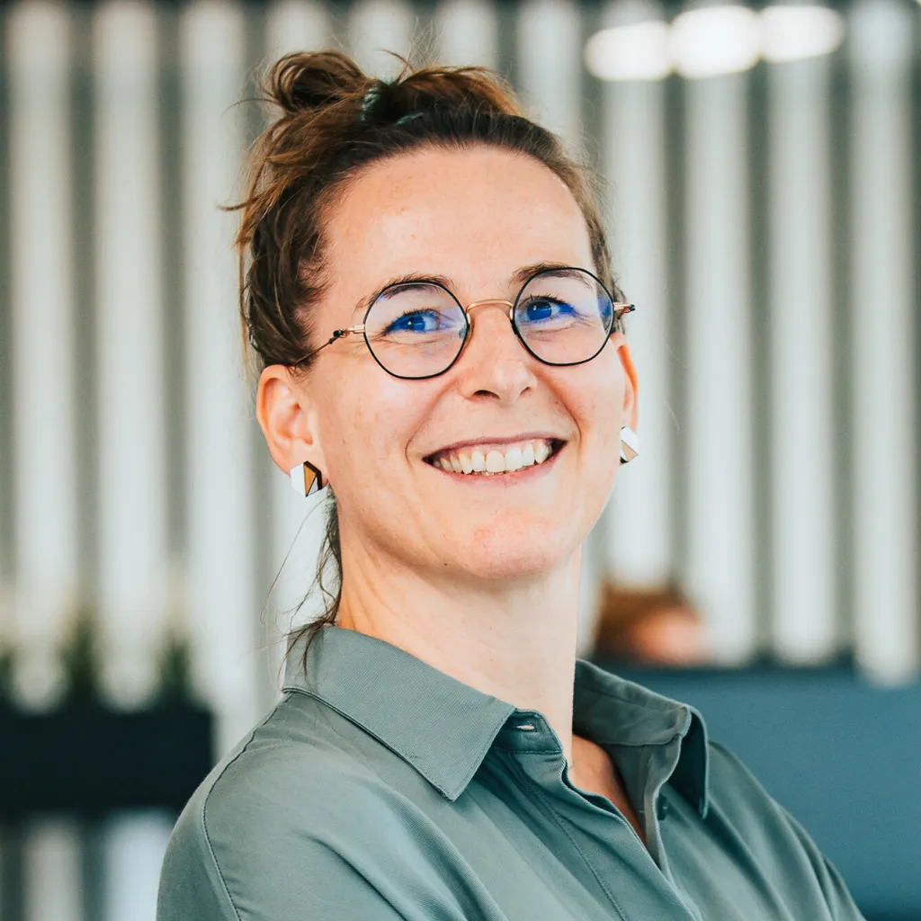 Smiling person with glasses and hair in a bun, wearing a green shirt in a bright, modern office.