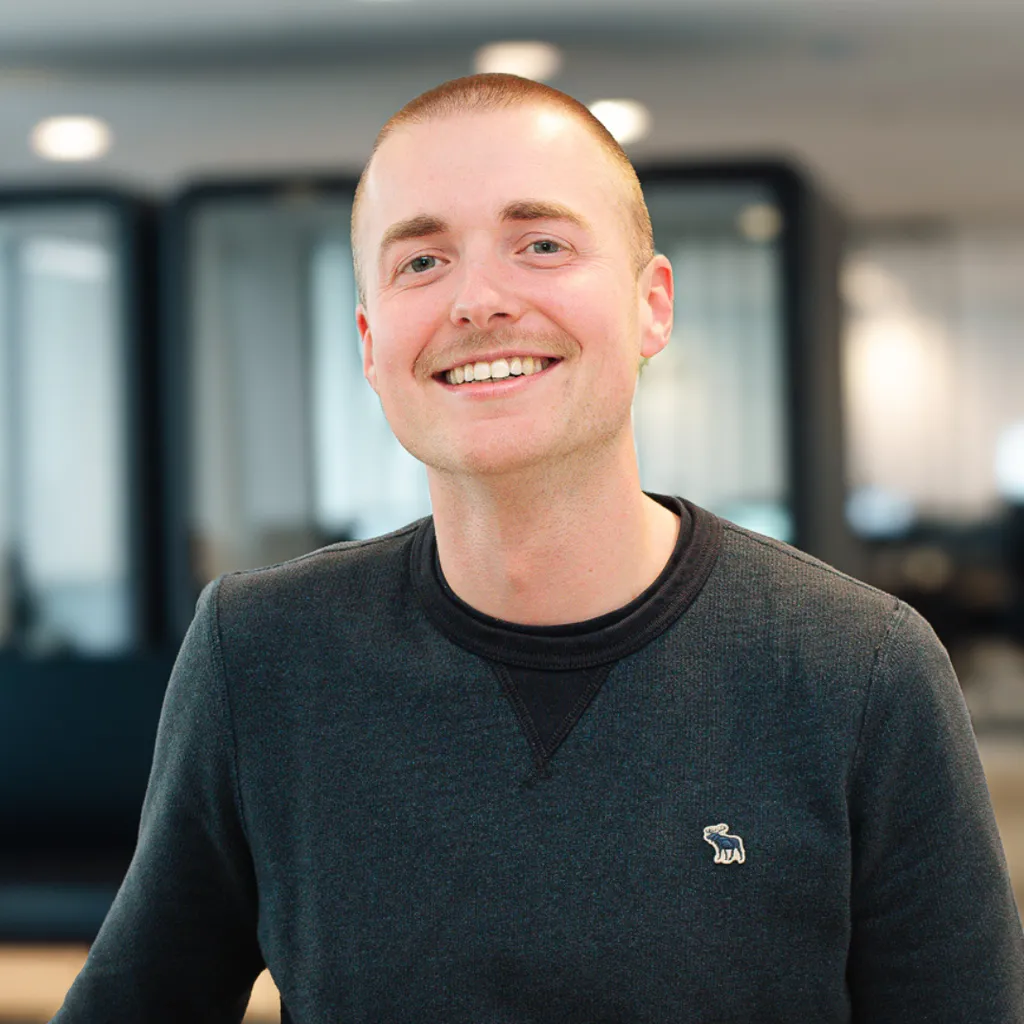 Smiling person in a dark sweater with a bear logo, standing in a modern office with blurred background.