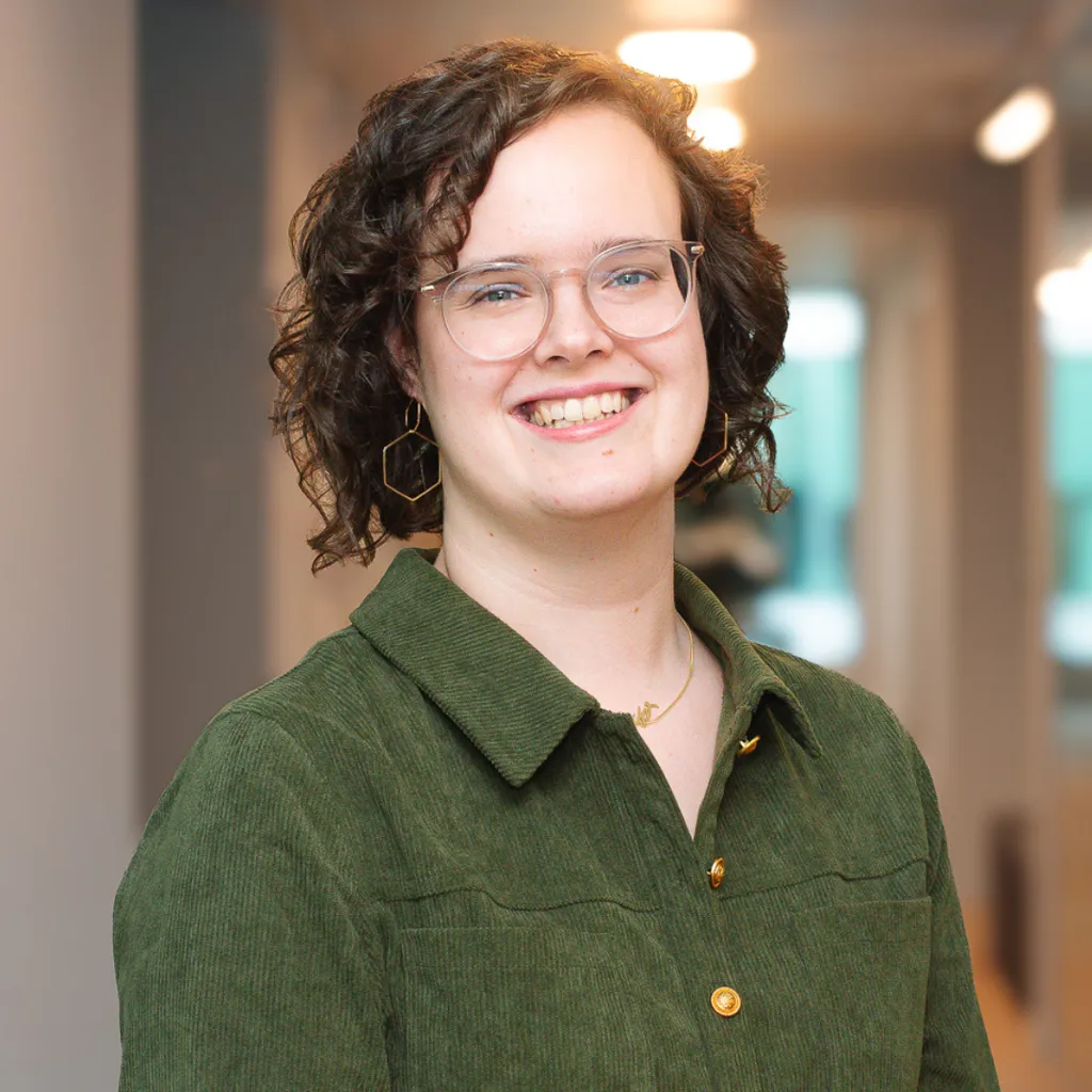 Smiling person with curly hair and glasses in a green shirt, standing in a bright hallway with blurred lights in the background.
