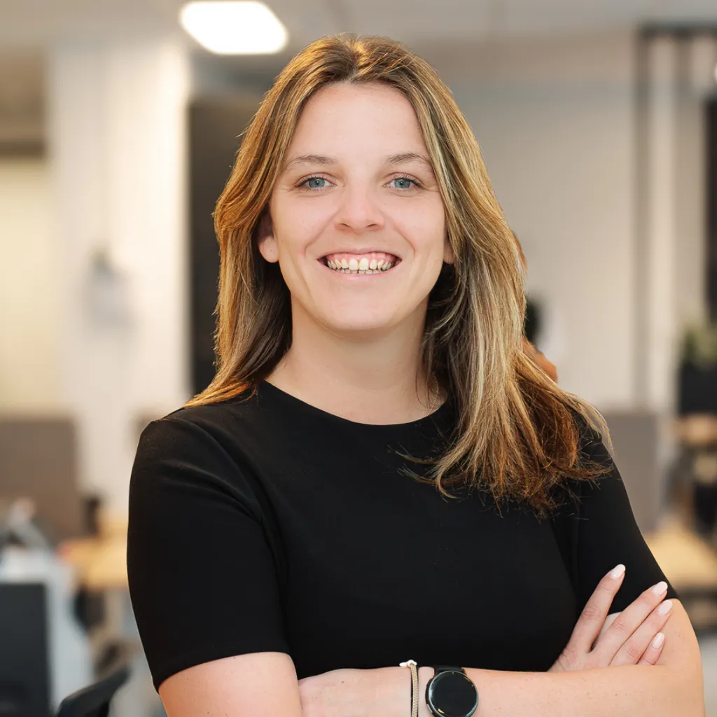 Smiling person with long hair in a black shirt, standing in a modern office setting with blurred background.
