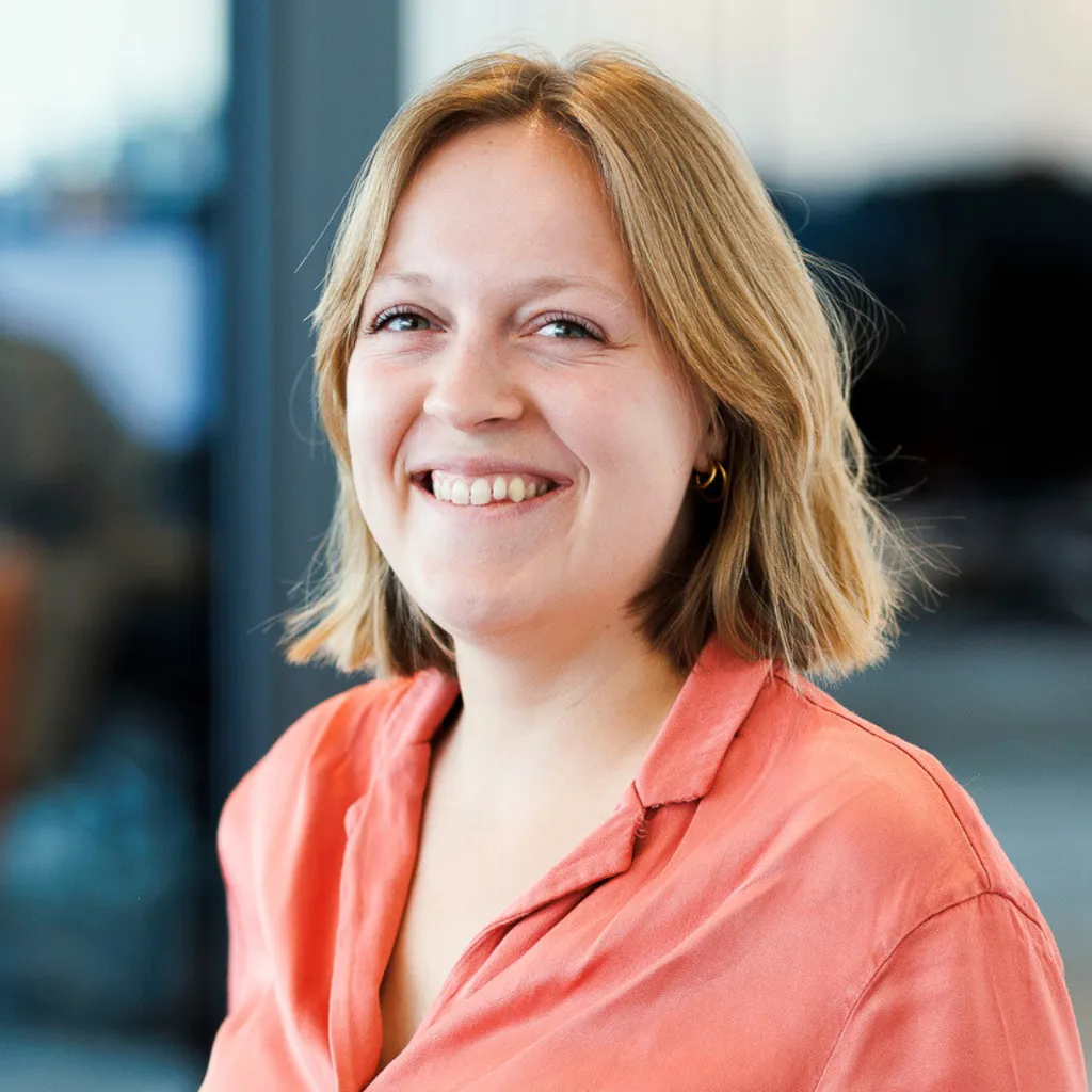 Smiling person with short blond hair and coral shirt standing indoors in a bright, modern office setting.