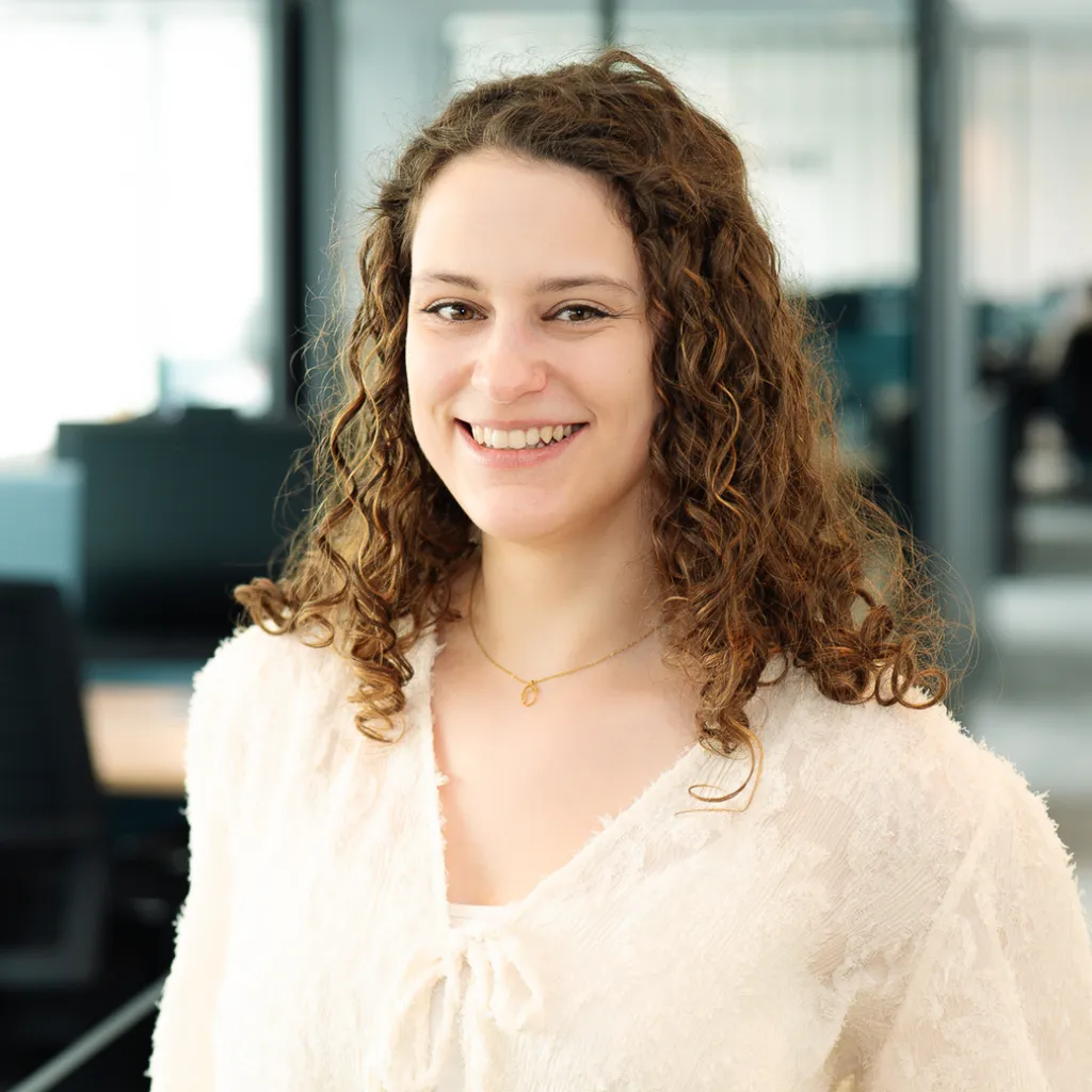 Smiling woman with curly hair in a light blouse standing in a bright modern office.