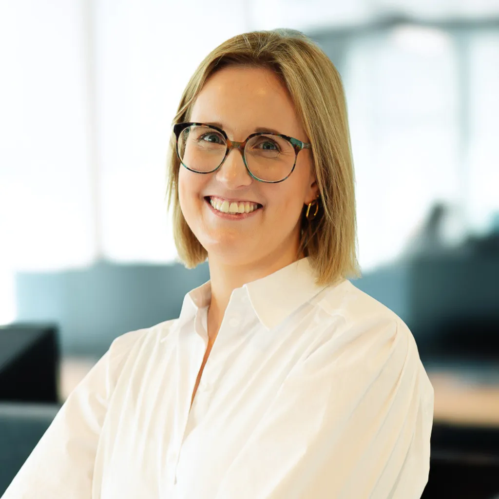 Smiling woman with short blond hair and glasses in a white shirt, posing in a bright modern office setting.