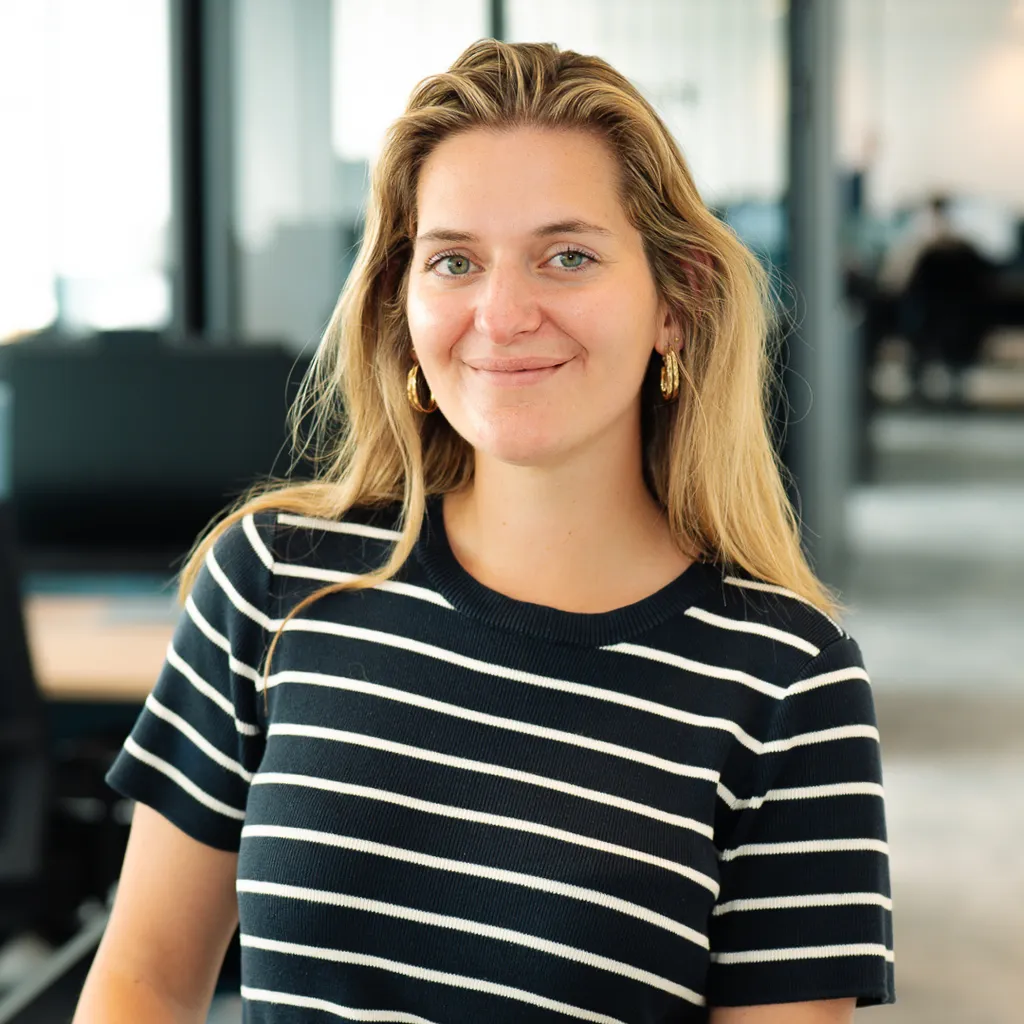Smiling woman in a striped shirt standing in a modern office with desks and coworkers in the blurred background.