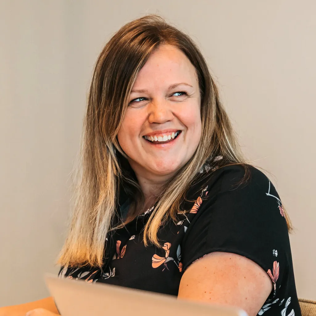 Smiling woman with long blonde hair in a floral top, seated indoors and looking to the side.