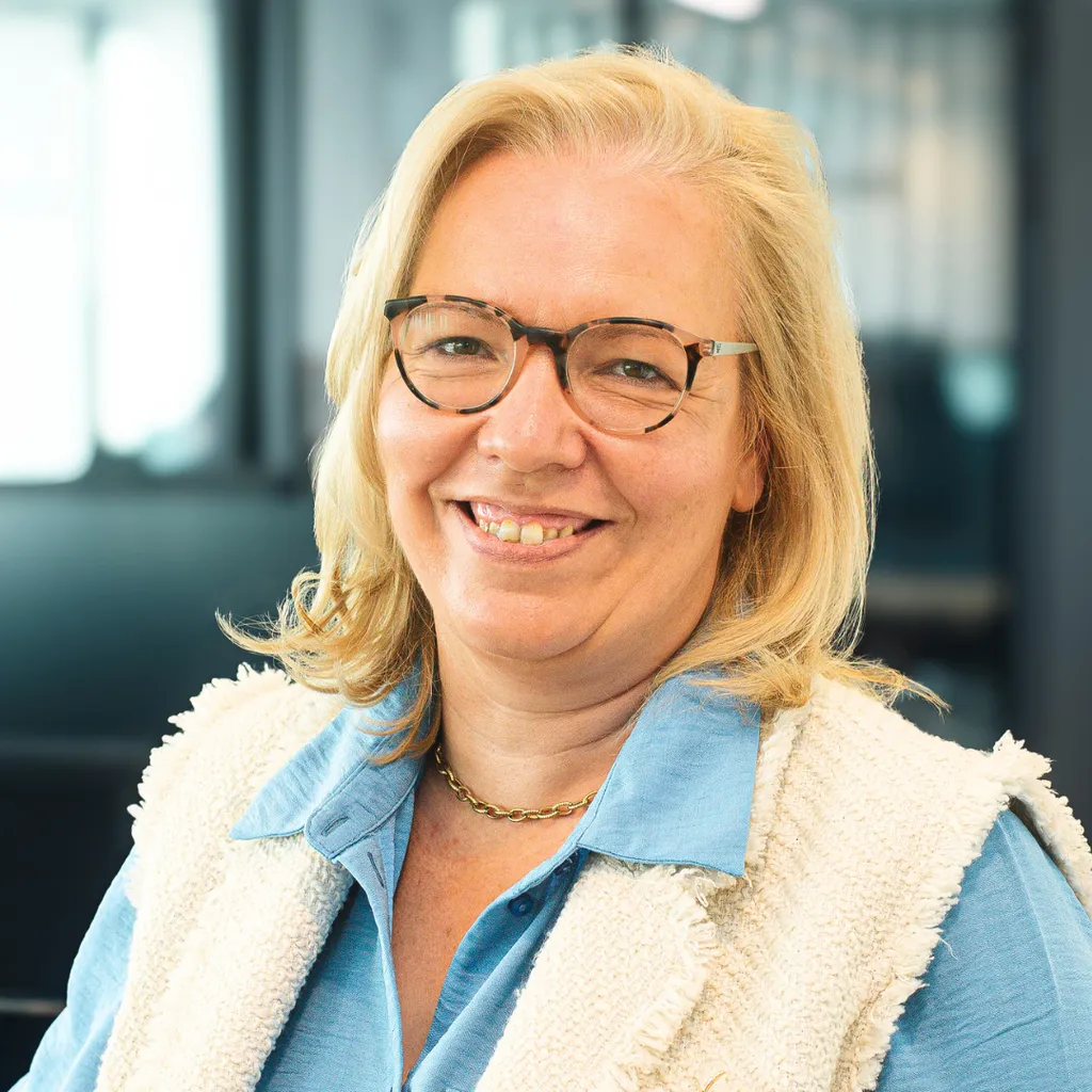 Smiling middle-aged woman with glasses and blonde hair in a light blue shirt and cream vest, standing in a modern office setting.