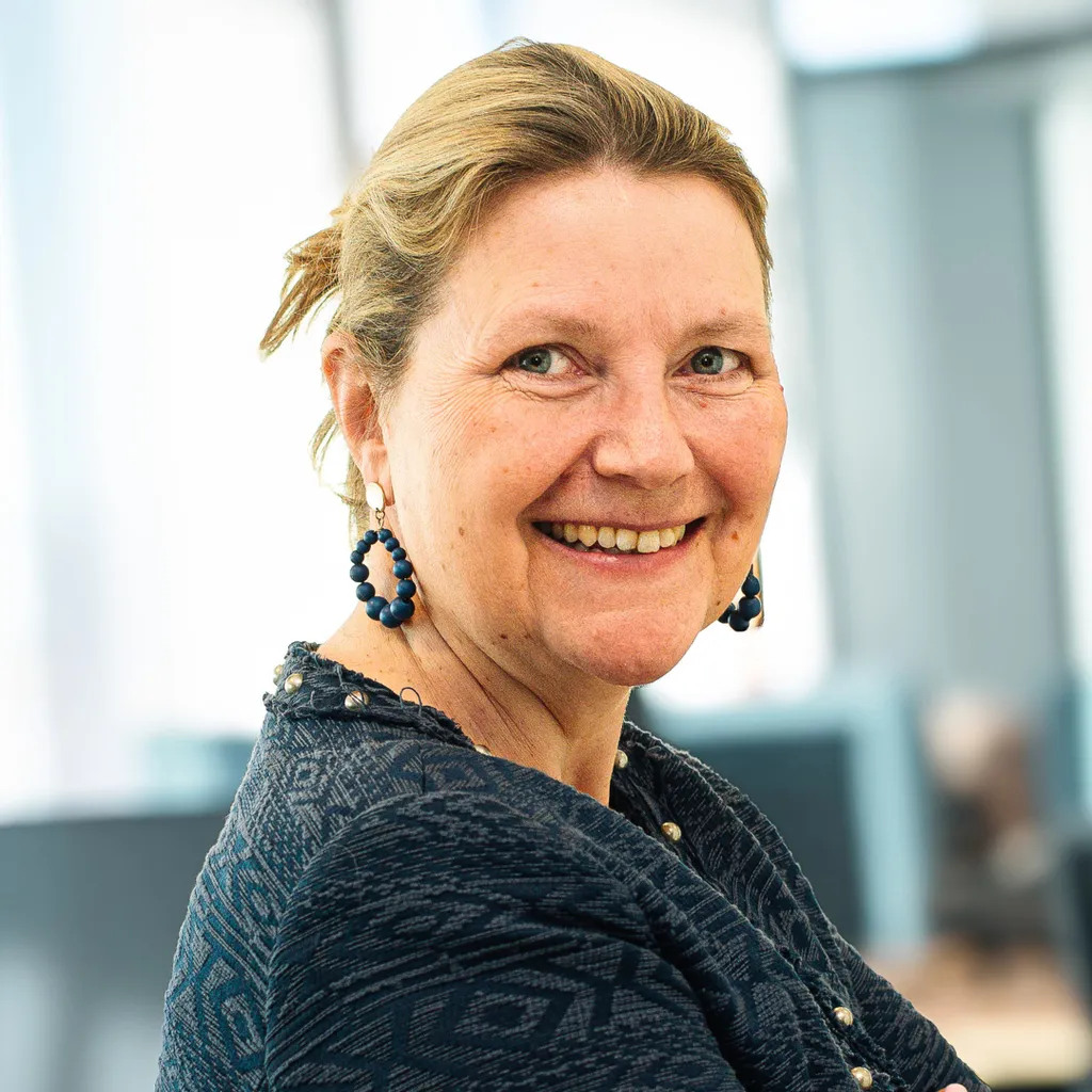 Middle-aged woman smiling at the camera in a bright office, wearing a patterned navy blazer and beaded earrings.