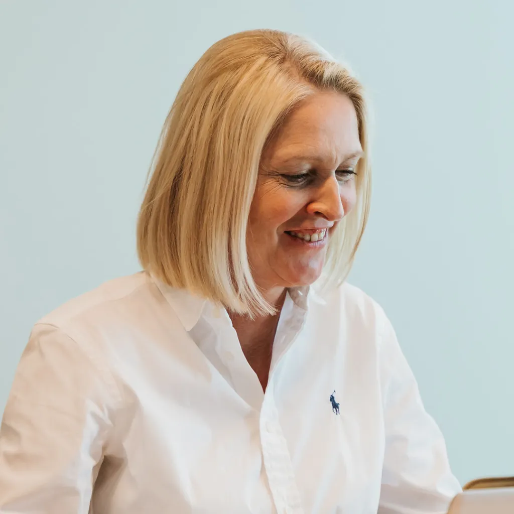 Smiling blonde woman in a white button-up shirt looking down, seated against a light blue background.
