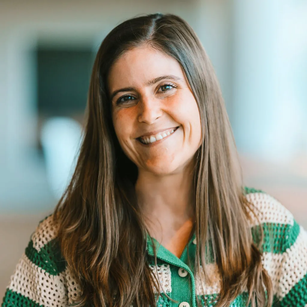 Smiling woman with long brown hair, wearing a green and beige striped sweater, stands indoors with a blurred background.