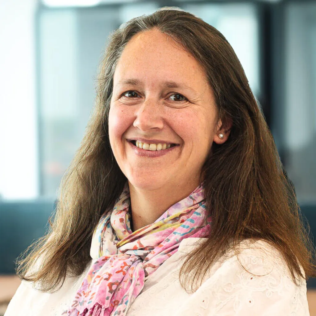Middle‑aged woman with long brown hair smiling at the camera, wearing a white top and patterned scarf in a bright office setting.
