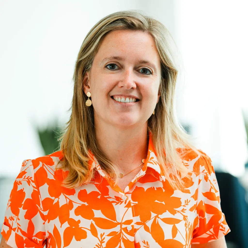 Smiling woman with long blonde hair wearing an orange floral shirt, standing indoors with a blurred background.