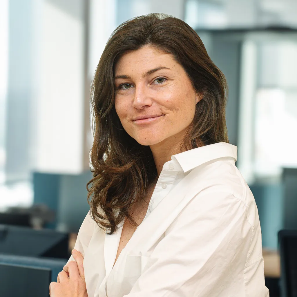 Woman with long brown hair in a white shirt smiles confidently in a modern, bright office setting.