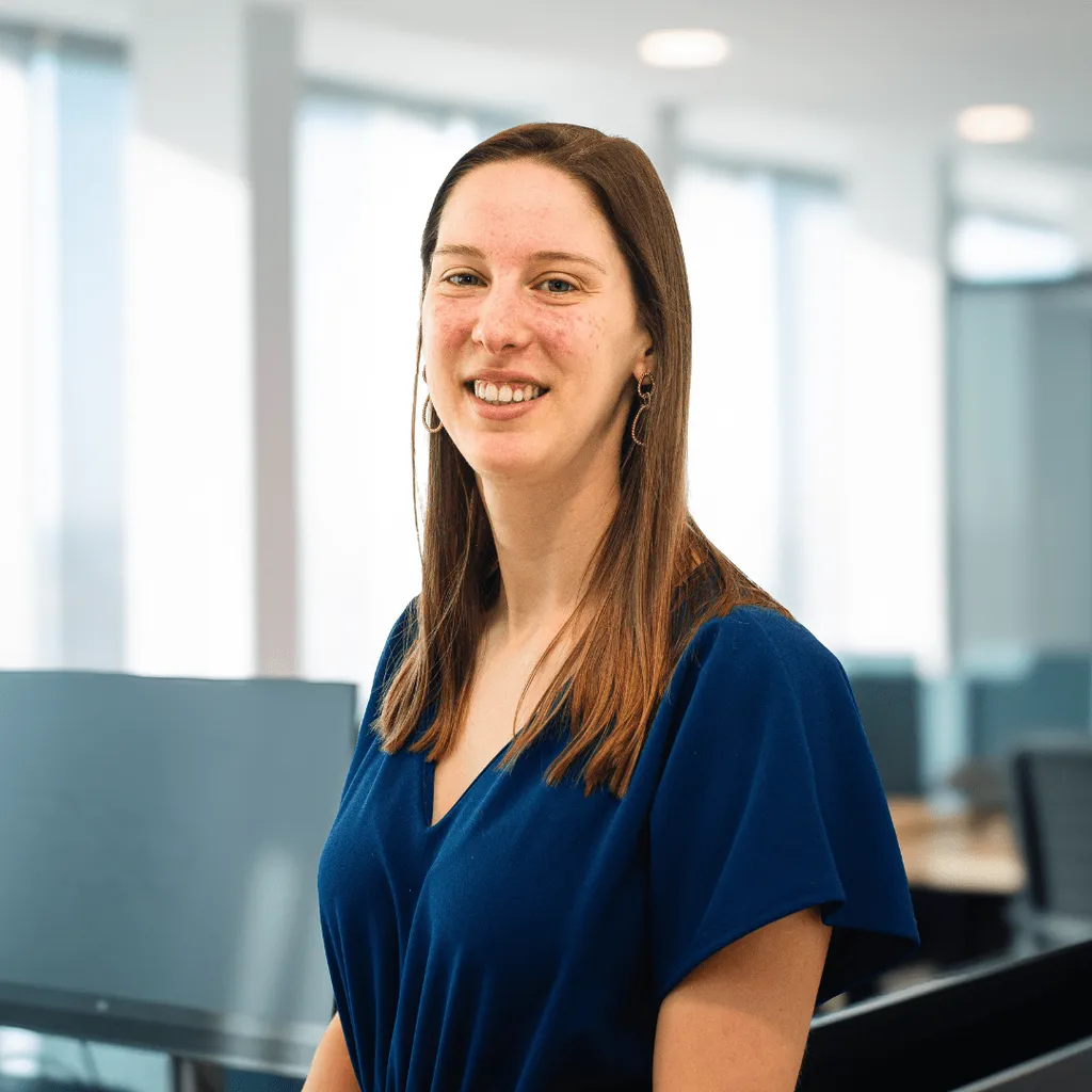 A smiling woman in an office environment, sitting at her desk with a computer and various office items.