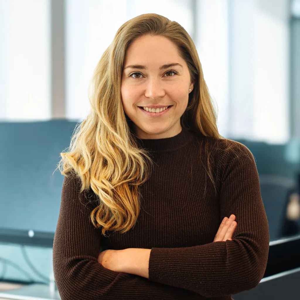 Smiling woman with long blonde hair, wearing a dark sweater, stands with arms crossed in a modern office setting.