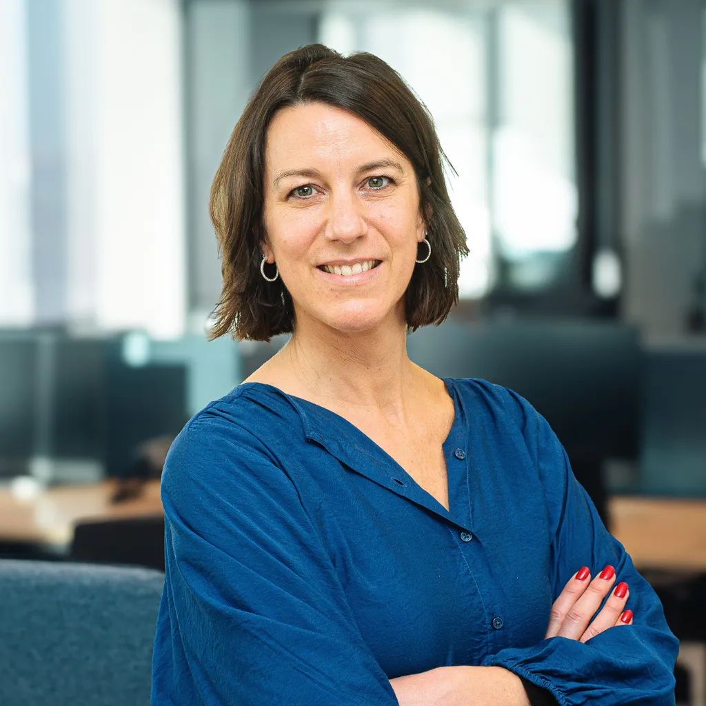 A woman with shoulder-length brown hair, wearing a blue blouse, stands confidently with arms crossed in a modern office setting.