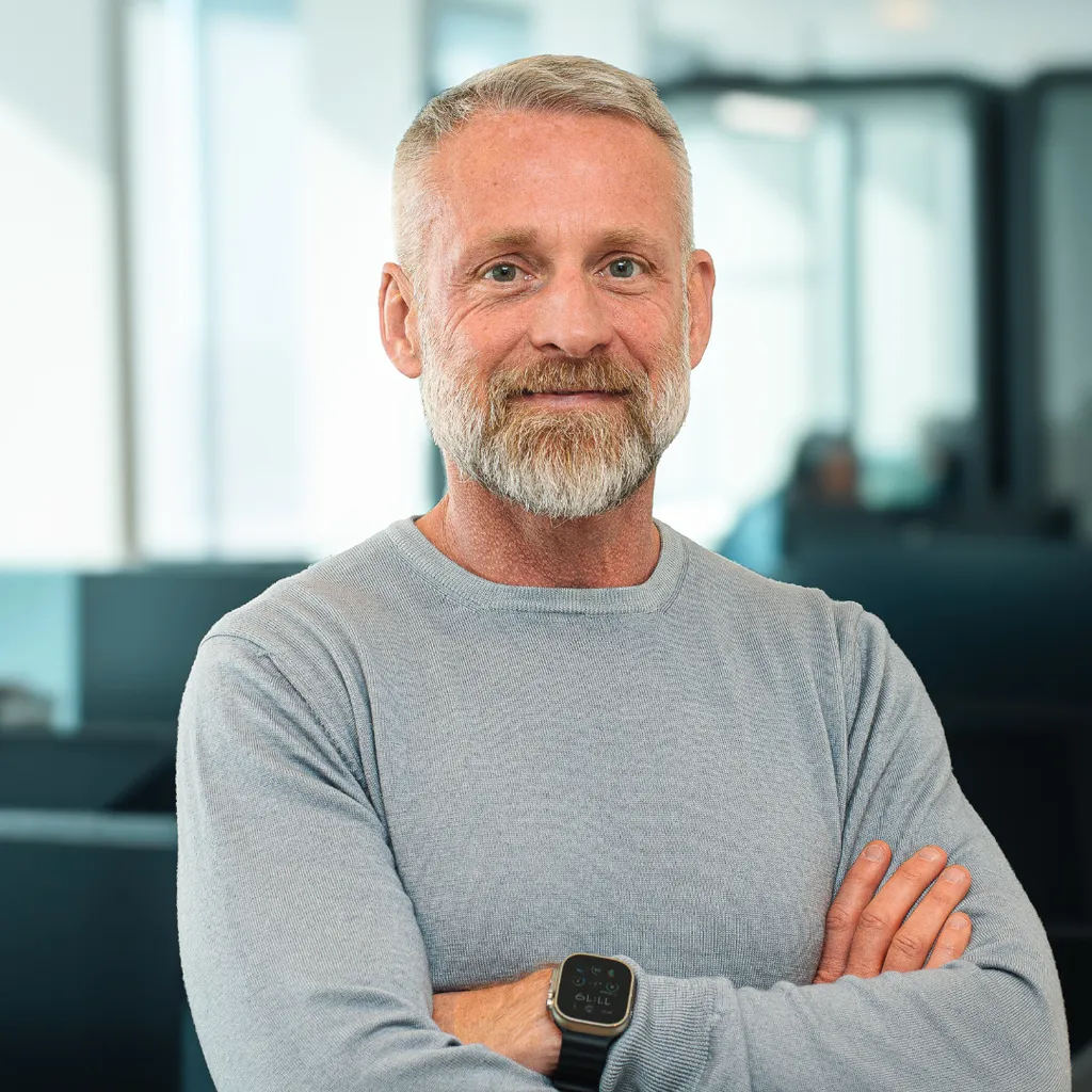 Man with gray hair and beard, wearing a light gray sweater and smartwatch, standing with arms crossed in a modern office setting.