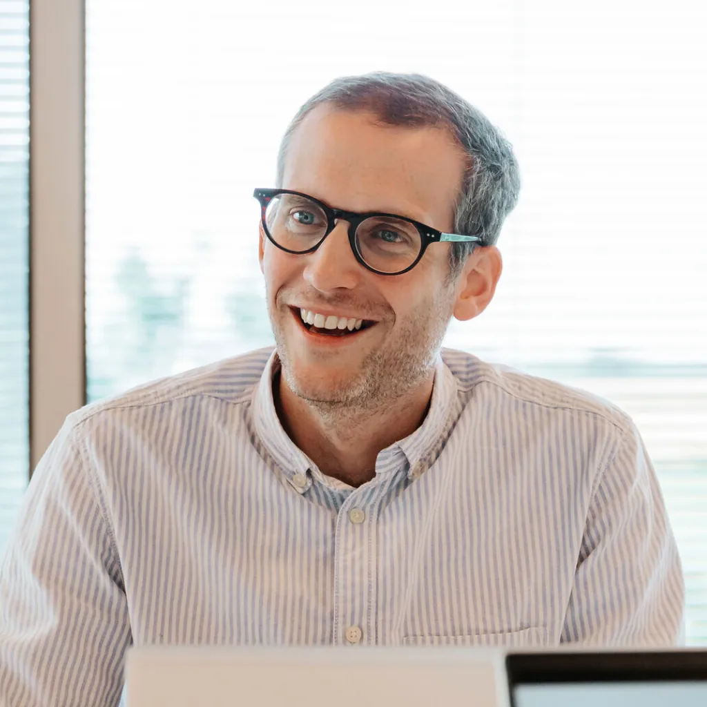 Smiling man with glasses and a striped shirt sits indoors, with blinds and a window in the background.