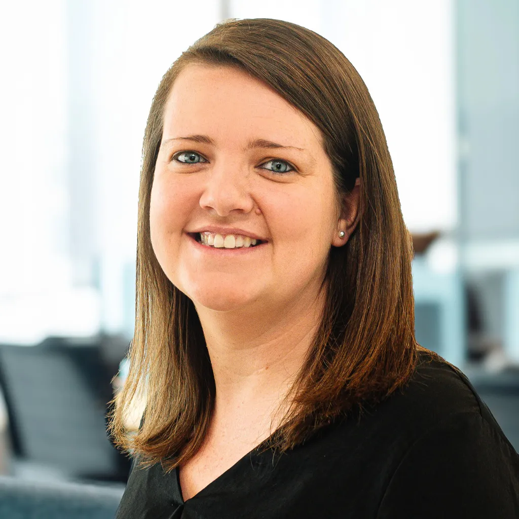 Smiling woman with straight brown hair in a black top, posing in a bright modern office setting.