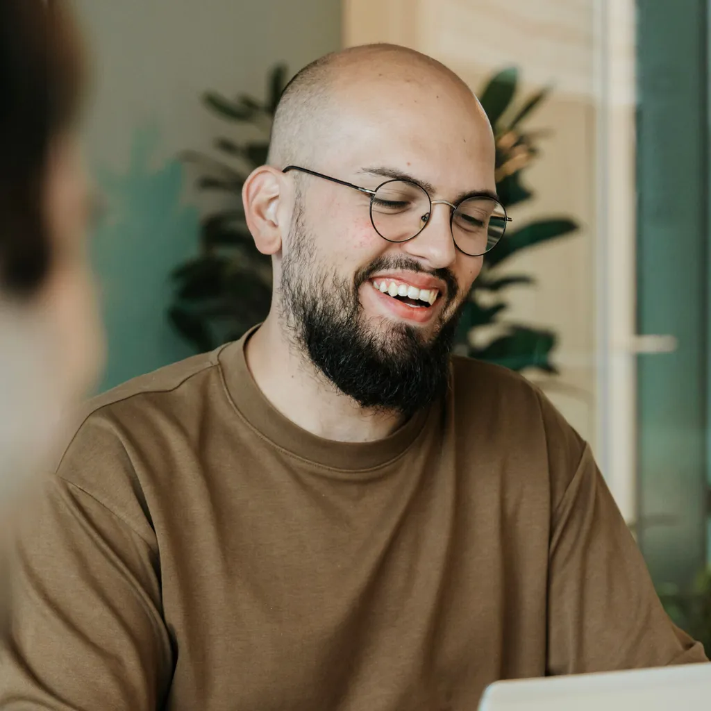 A bearded man with glasses smiles while sitting at a table, wearing a brown shirt. Blurred background with plants visible.