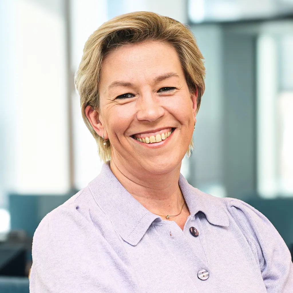 Smiling middle‑aged woman in a light purple blouse stands in a bright modern office, looking confidently at the camera.