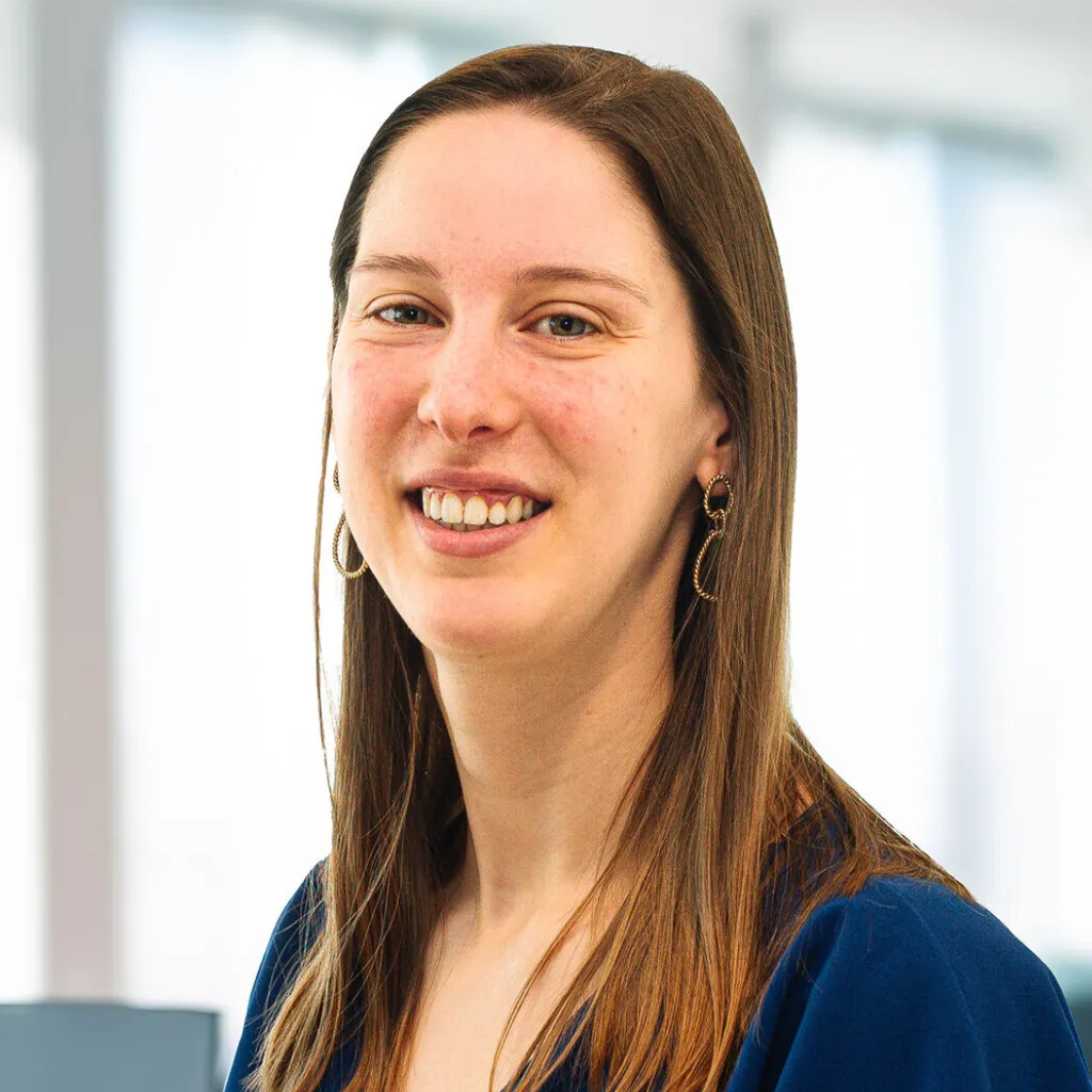 Smiling person with long straight hair wearing a blue top and dangly earrings, standing in a bright office setting.