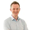 Smiling man in a light blue shirt with arms crossed, standing against a plain white background.