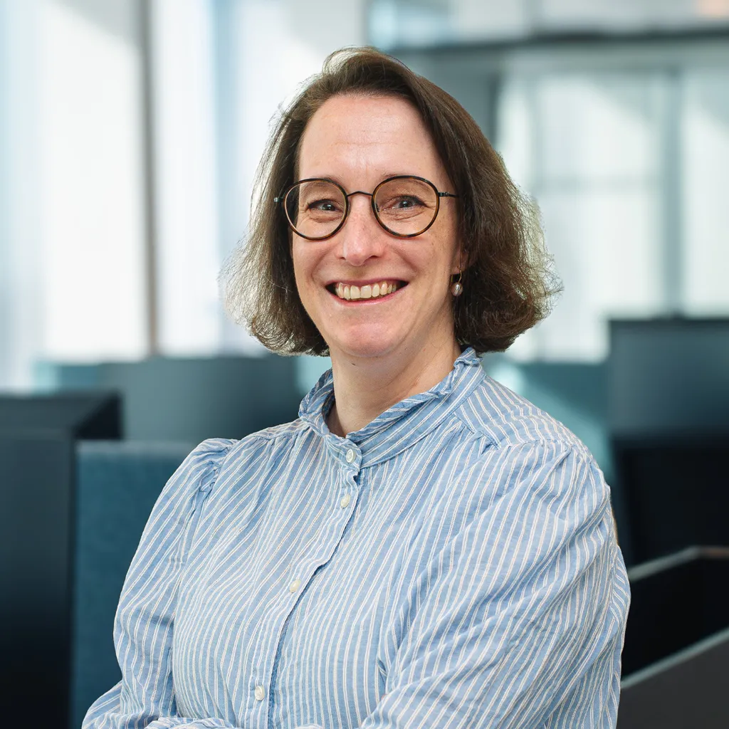 A woman with glasses and a striped shirt smiles, standing in a modern office setting with large windows in the background.