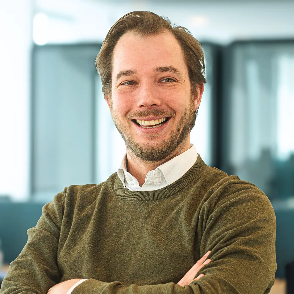Smiling man with light beard in sweater and collared shirt, standing with arms crossed in a bright modern office.