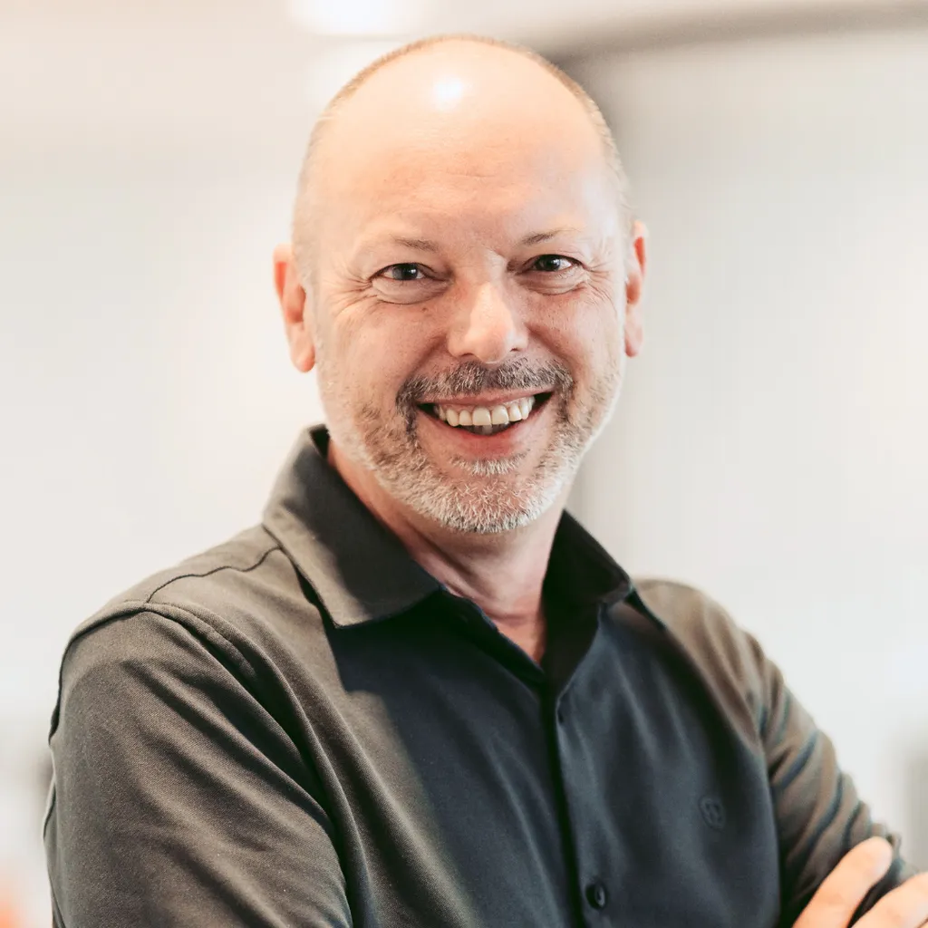 Smiling middle‑aged man with a shaved head and gray beard, wearing a dark shirt, standing in a bright indoor setting.