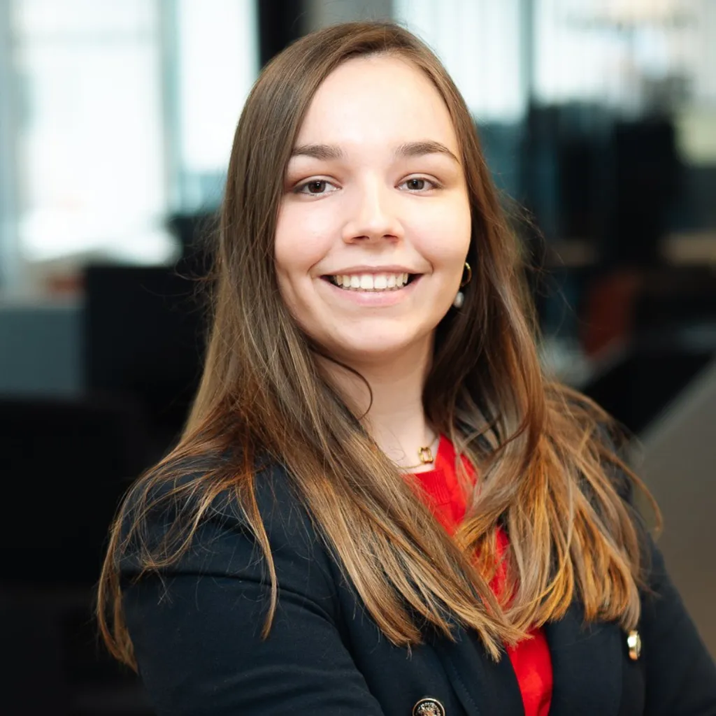 Smiling young woman with long brown hair in a blazer and red top, posing confidently in a modern office setting.