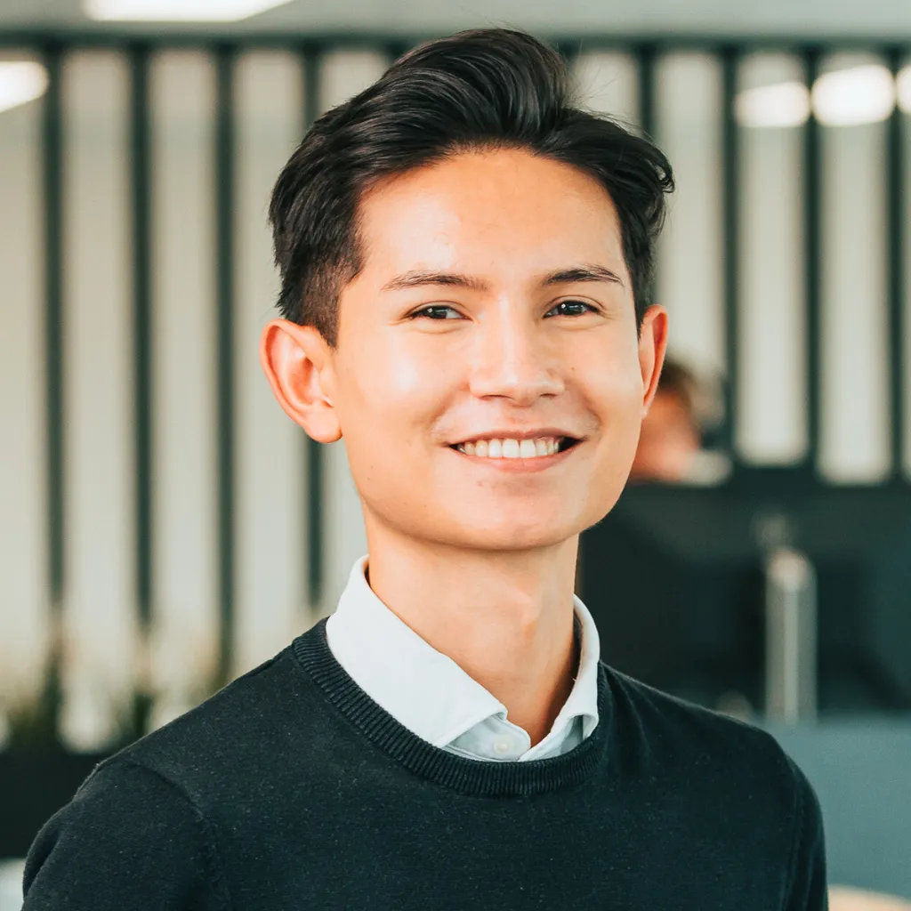 Smiling young professional in a dark sweater and collared shirt, standing in a modern office with blurred background.