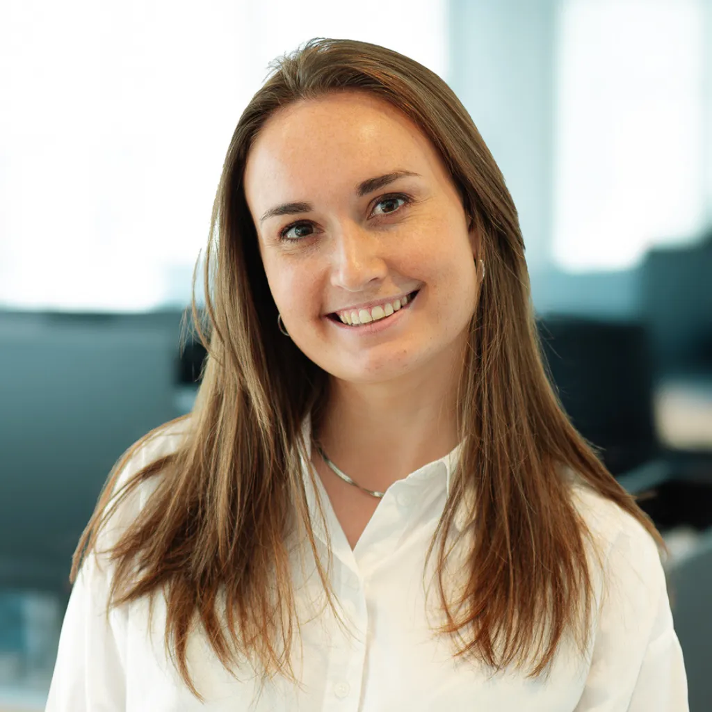 Young woman with long brown hair smiling in a bright office, wearing a white shirt and silver necklace.