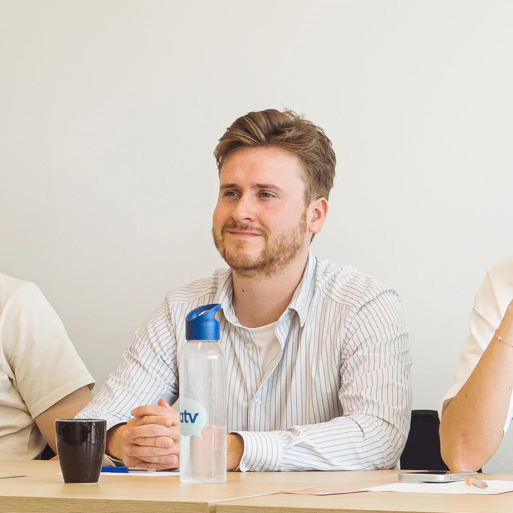 Young man in a striped shirt sits at a desk, listening attentively with a water bottle and coffee cup in front of him.