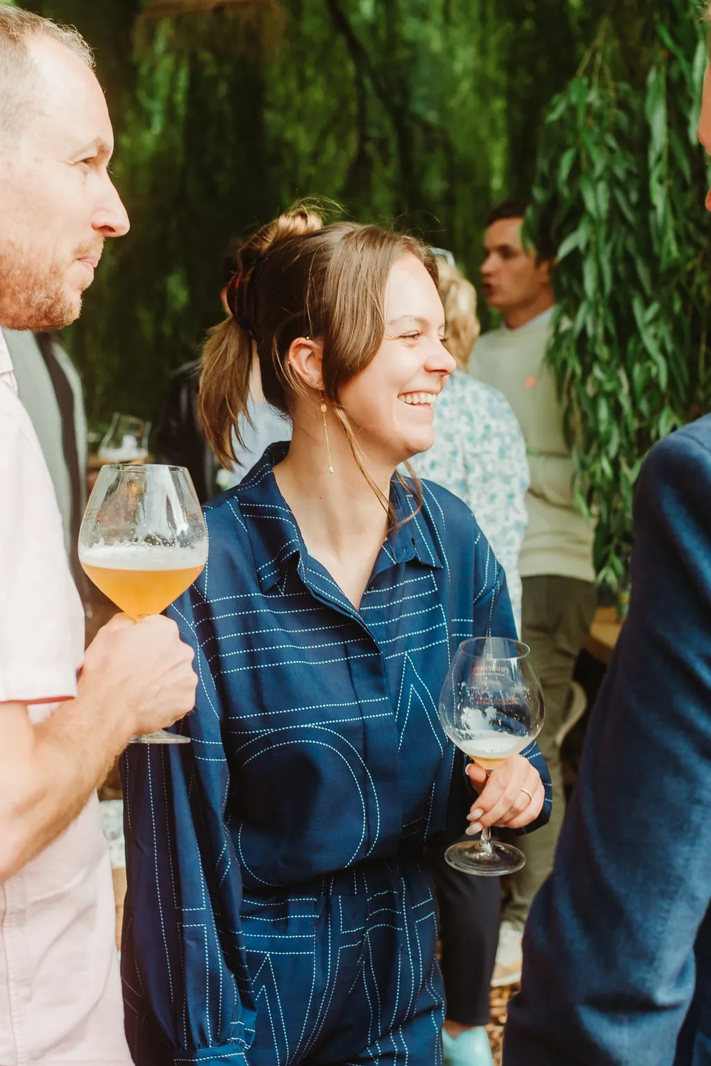 Woman in a blue outfit smiles while holding a wine glass at an outdoor gathering, surrounded by people and greenery.